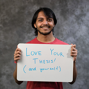 A young man holding up a sign saying: Love Your Thesis and yourself