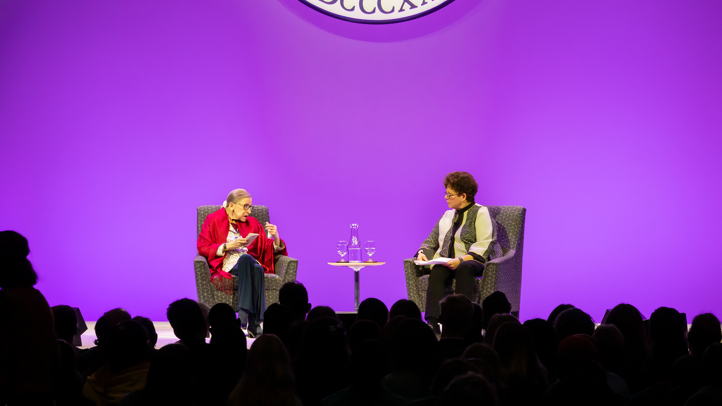 A photo of Ruth Bader Ginsburg speaking with President Biddy Martin on stage against a purple background