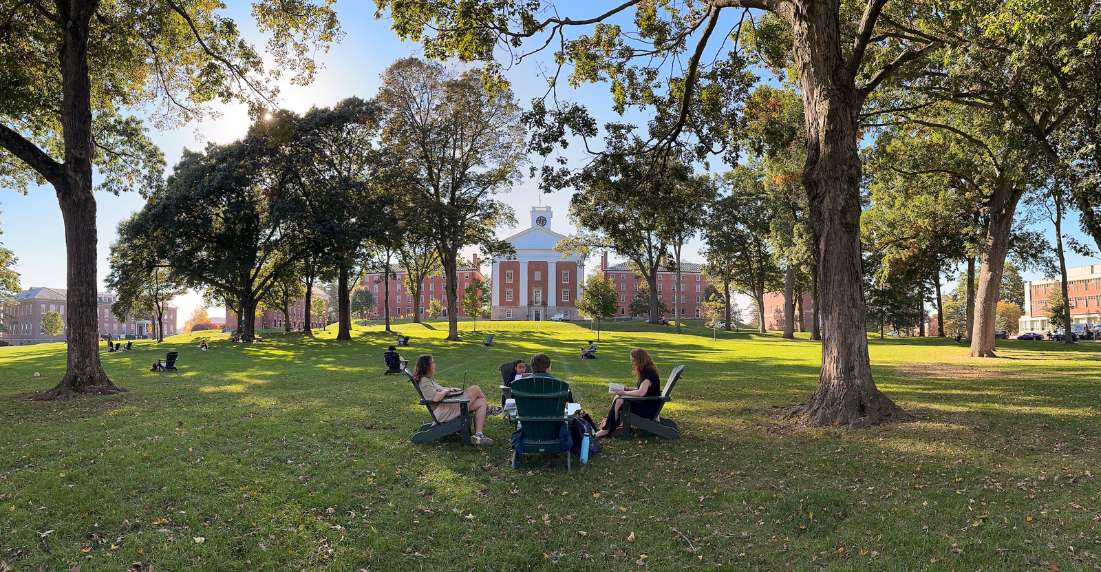 students sit in a group of Adirondack chairs on the quad with Johnson Chapel behind them