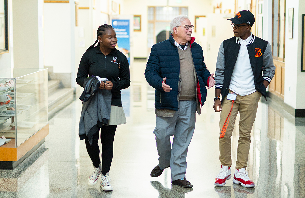 An older man walking with two students in a school hallway