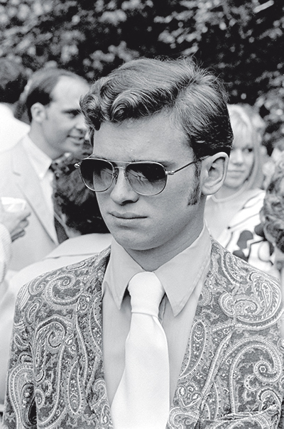A black and white photo of a 1970s young man in sunglasses and a paisley suit