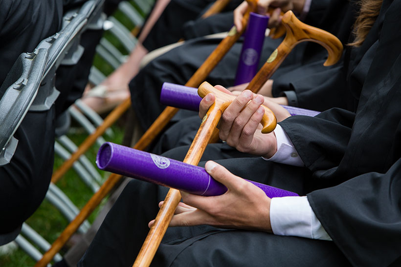 Students in commencement gowns holding canes and purple diploma tubes