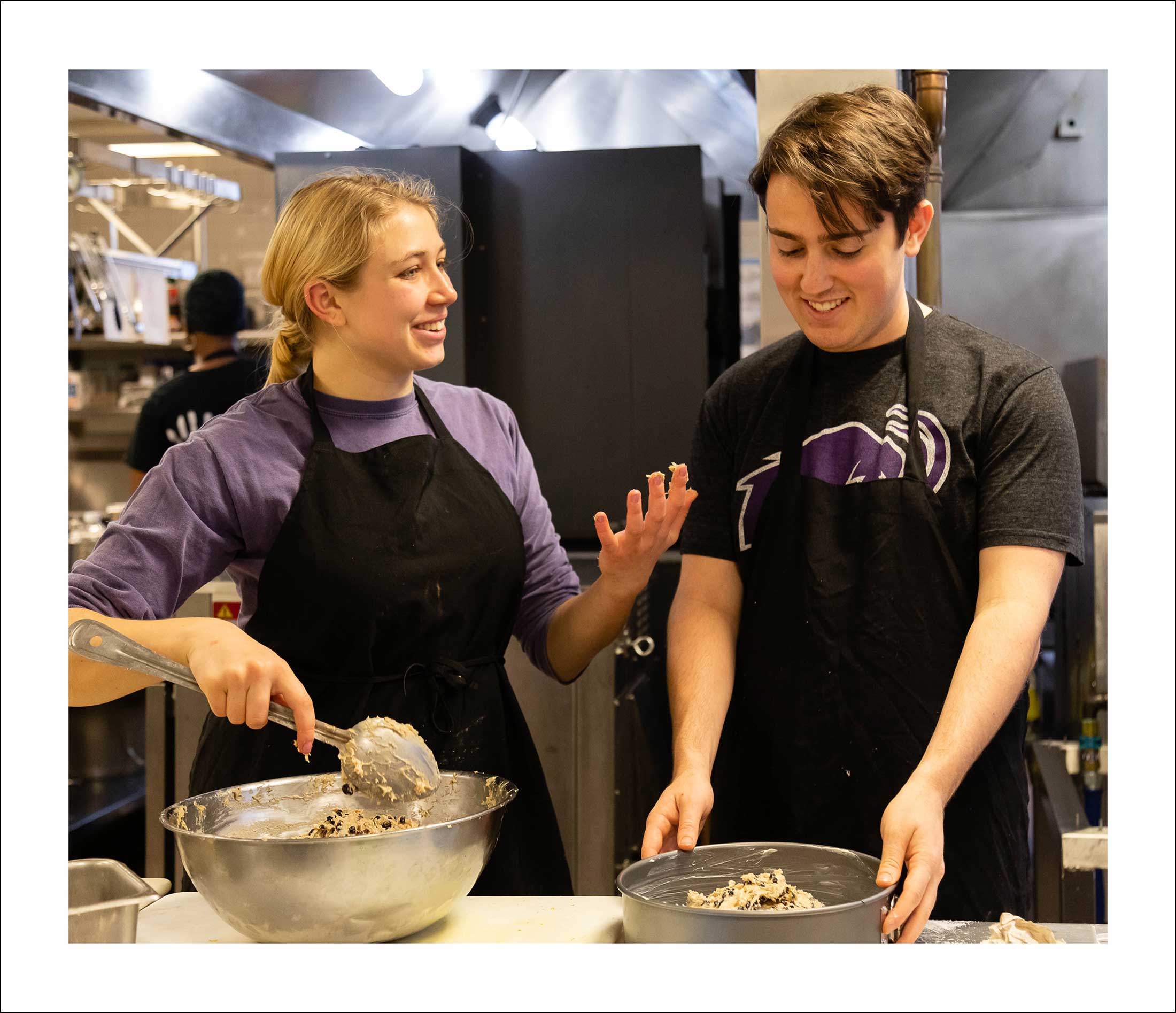 A young man and woman wearing black aprons stir ingrediants in large silver bowls