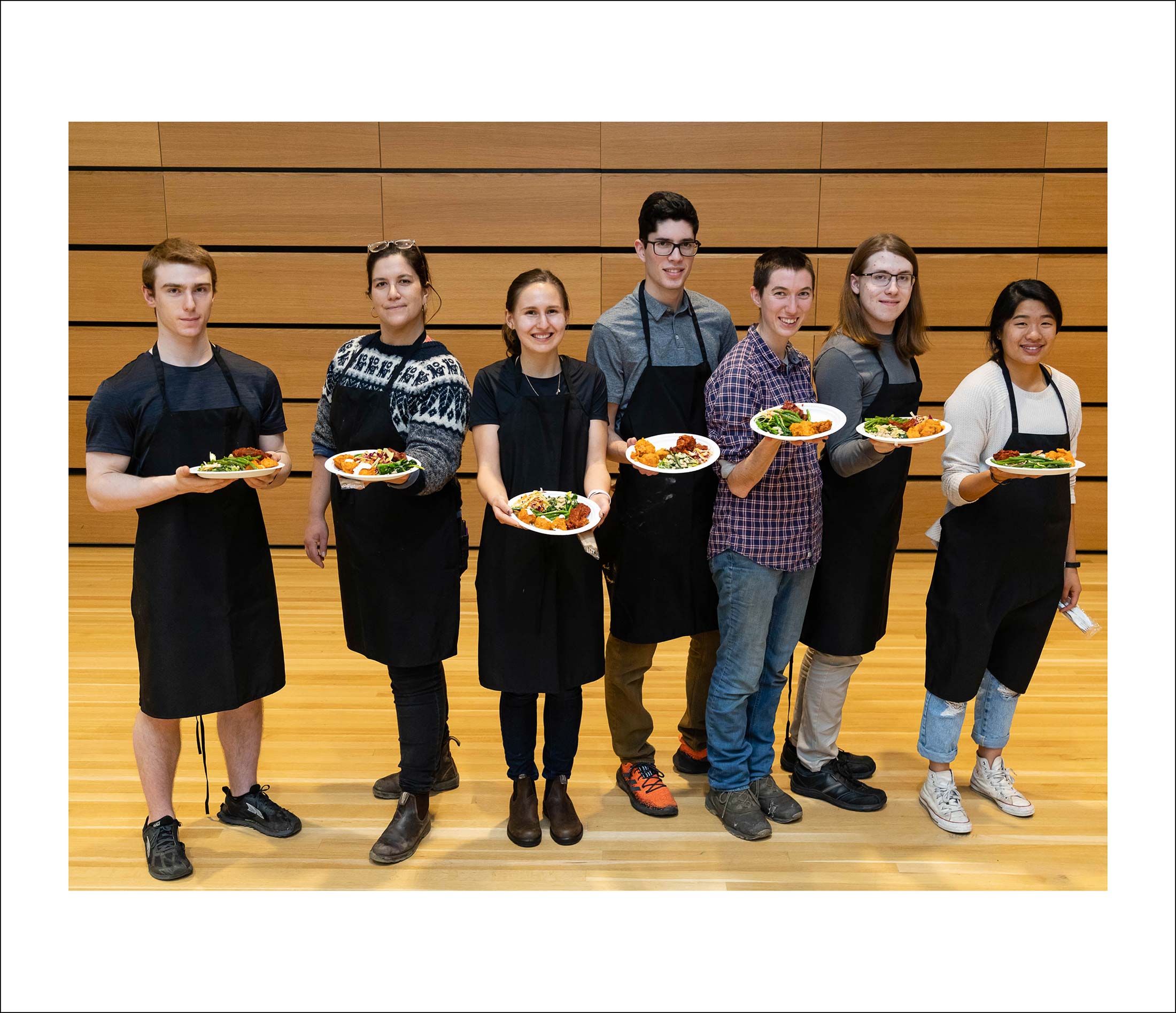 Seven people wearing black aprons, each holding a plateful of food