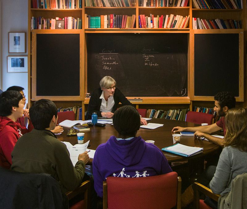 Philosophy professor sits at a table with her class of 6 students in a cozy room lined with book shelves