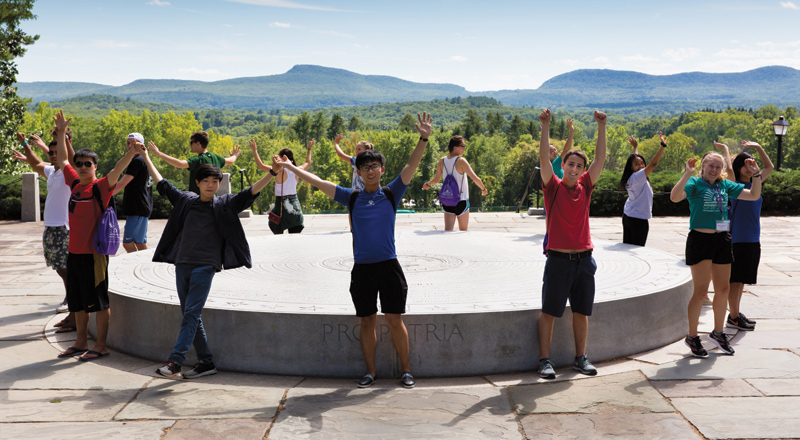 Students stand in a circle holding their hands, joined, up in the air, with a mountain range behind them